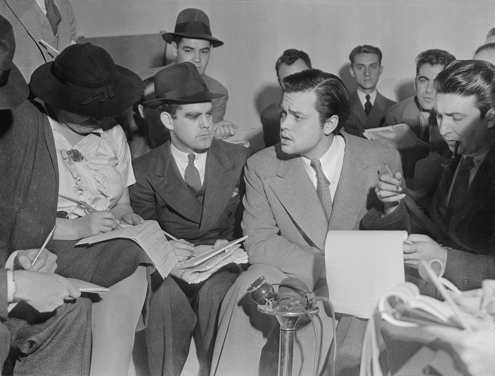 Photo: Orson Welles, in a light jacket and patterned tie, talks with a crowd of reporters who are writing on notepads, with a microphone in front of them.