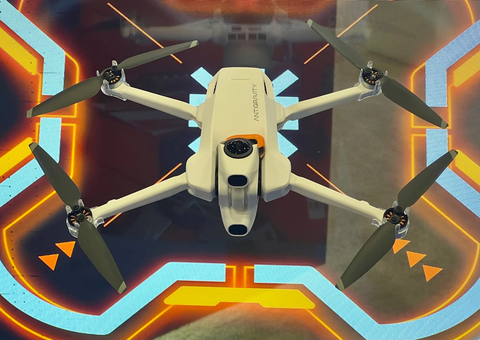A white Antigravity A1 drone with its four propellers unfolded, in front of a glowing orange and blue display.