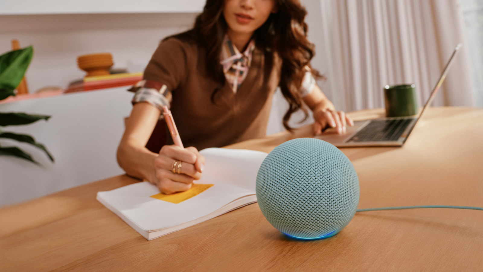 Photo: A blue Echo Dot sphere with a glowing light ring sits on a wooden desk while a woman writes in a notebook.