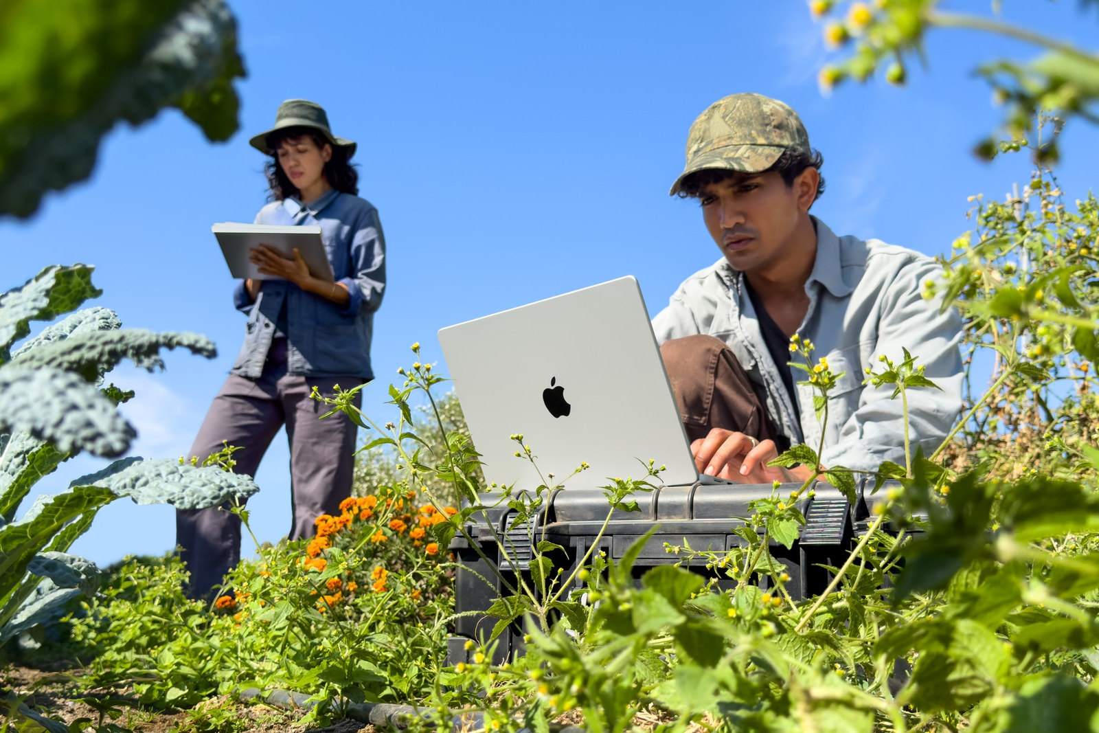 Photo: A man works on a silver MacBook Pro set on a case in a garden while a woman behind him holds an iPad.
