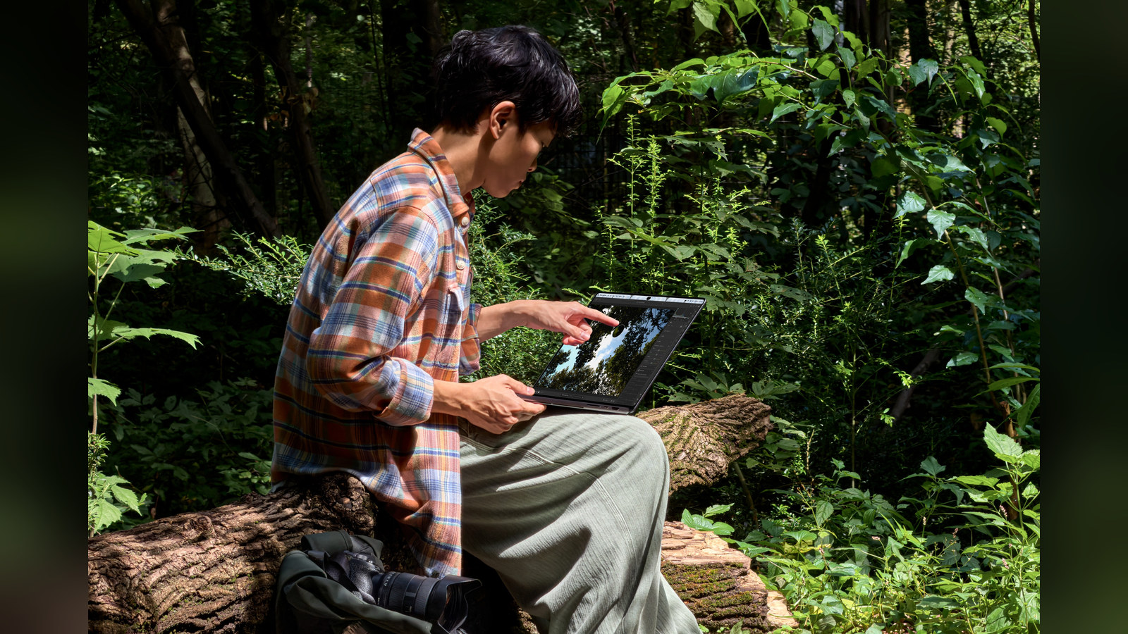 Photo: A person sitting on a log in a forest touches the screen of the MacBook Pro with PROLED XDR Display.