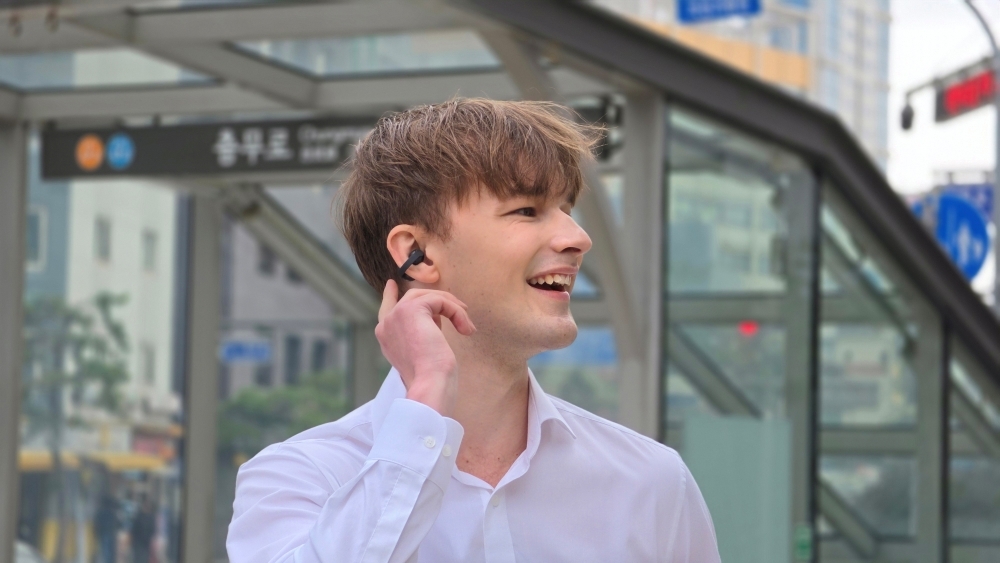 A man in a white shirt touching an earbud and smiling at the entrance to a subway station